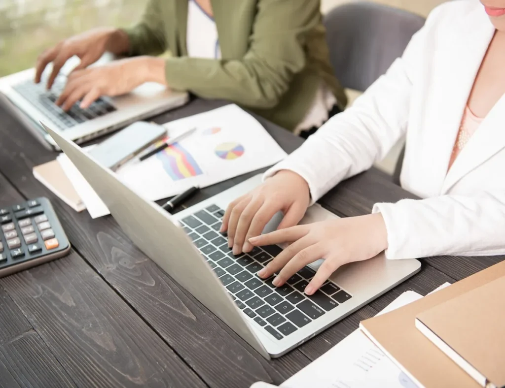 Two bookkeepers working at laptops with financial reports beside them